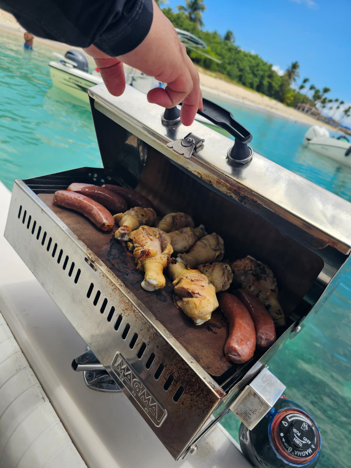 Chicken legs and sausages cooking on the onboard Magma marine grill with turquoise Caribbean water and beach in the background