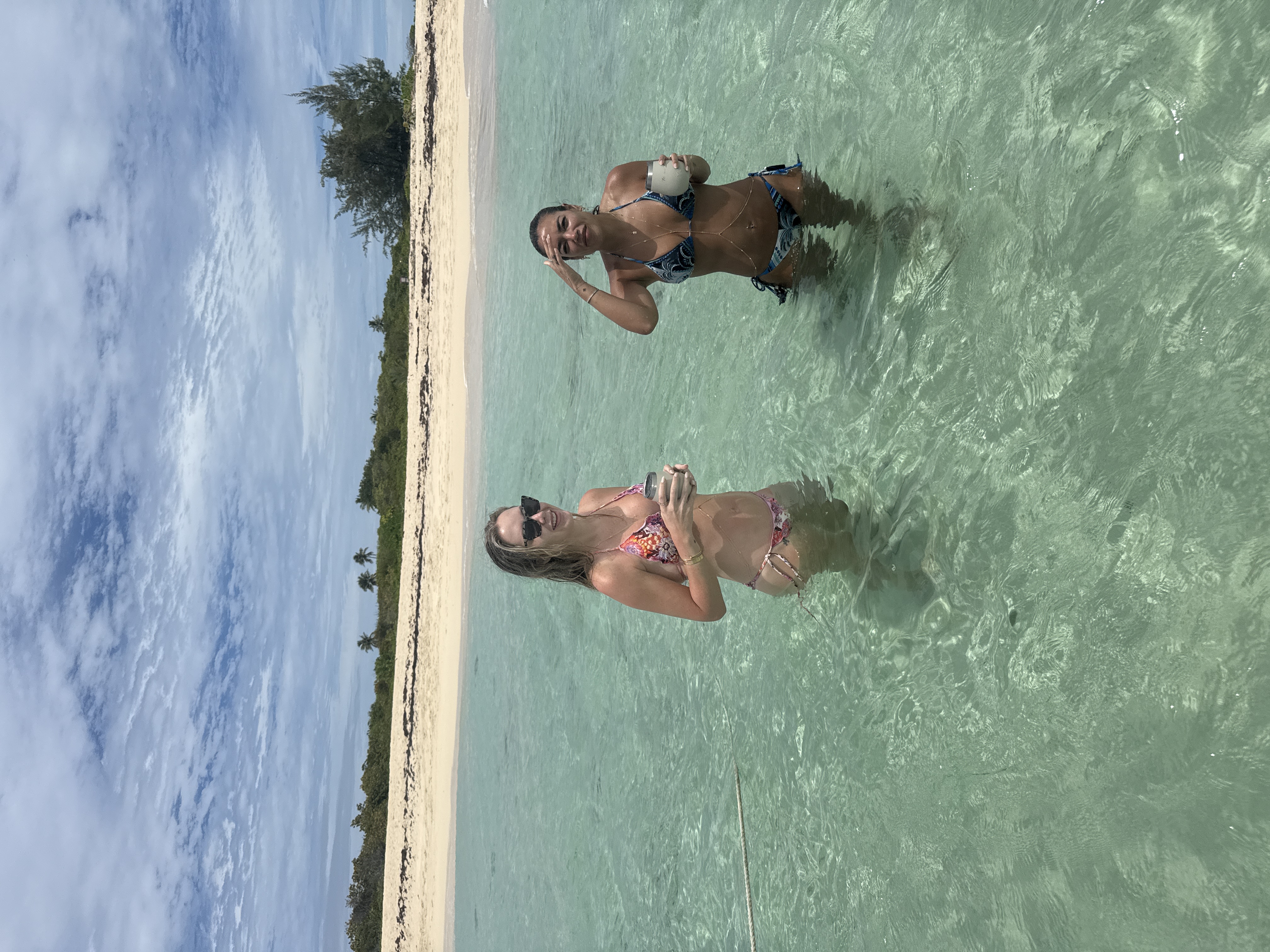Two guests in shallow turquoise water with drinks at the sandbar