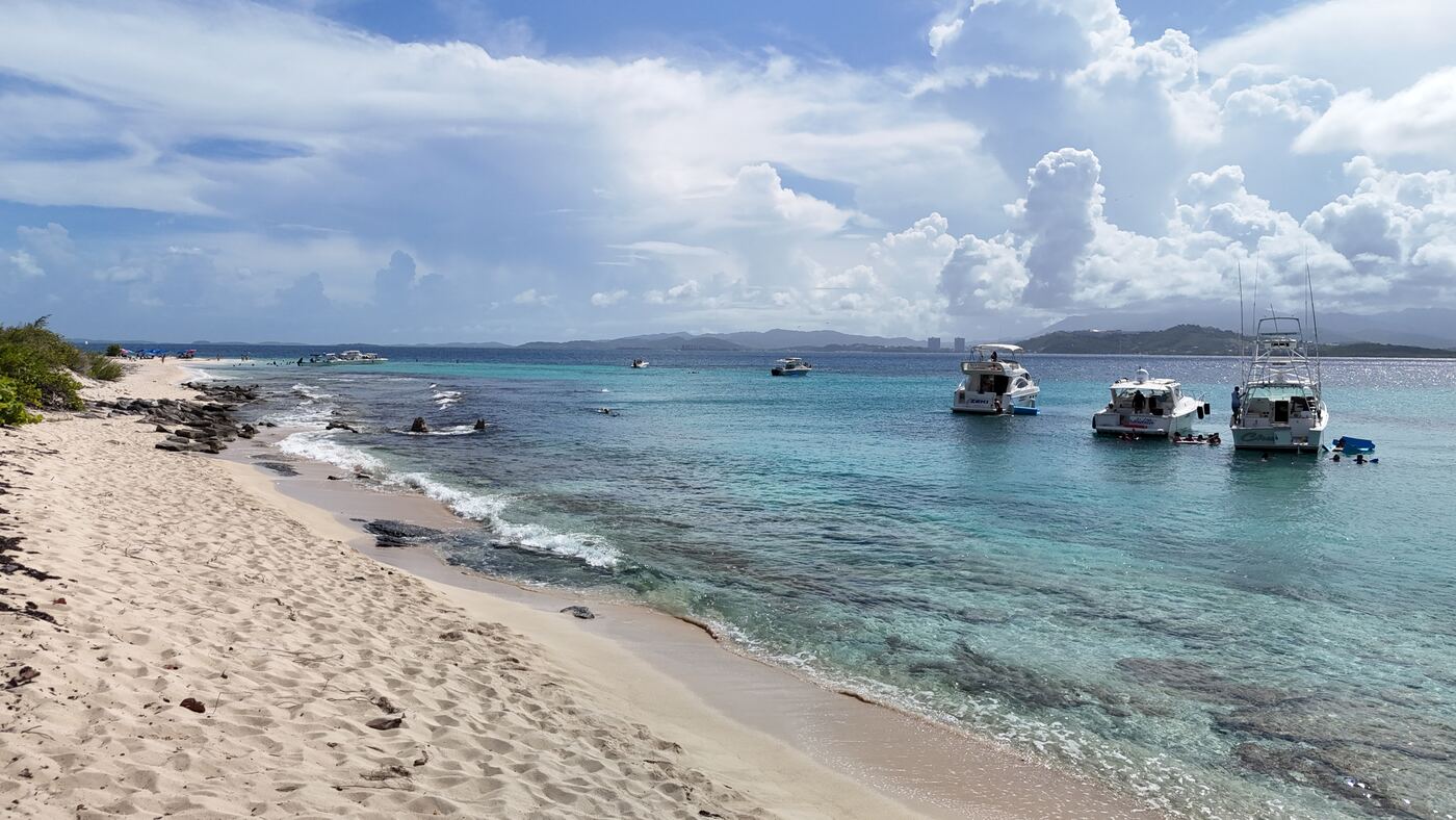 Wide view along the Icacos shoreline with boats anchored offshore, guests snorkelling, and the mountains of Puerto Rico across the water from Fajardo