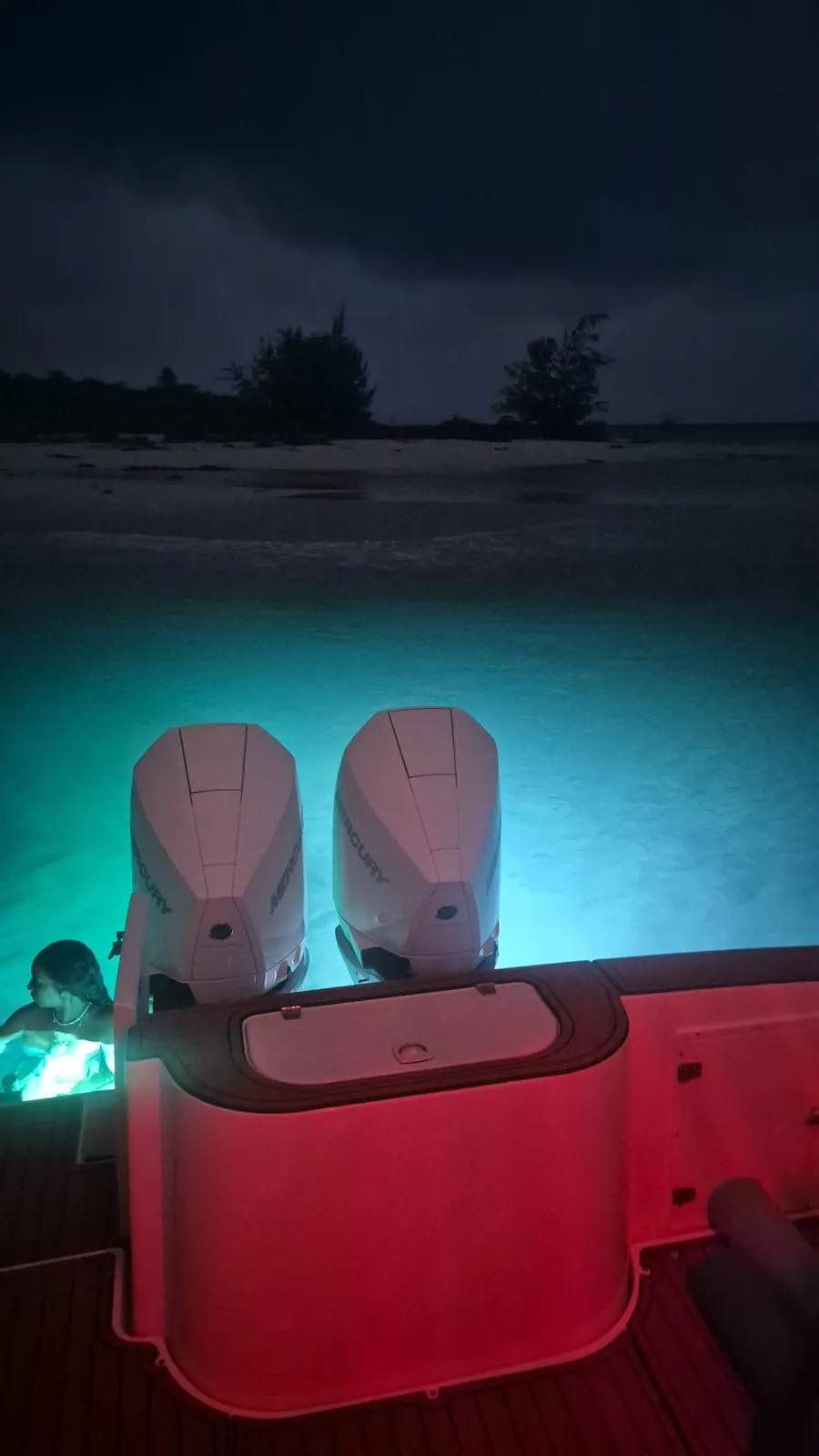 Stern of the Contender 38 at night off Palomino Island, twin Mercury outboards above turquoise water lit by the underwater lights, with the dark treeline of the beach behind