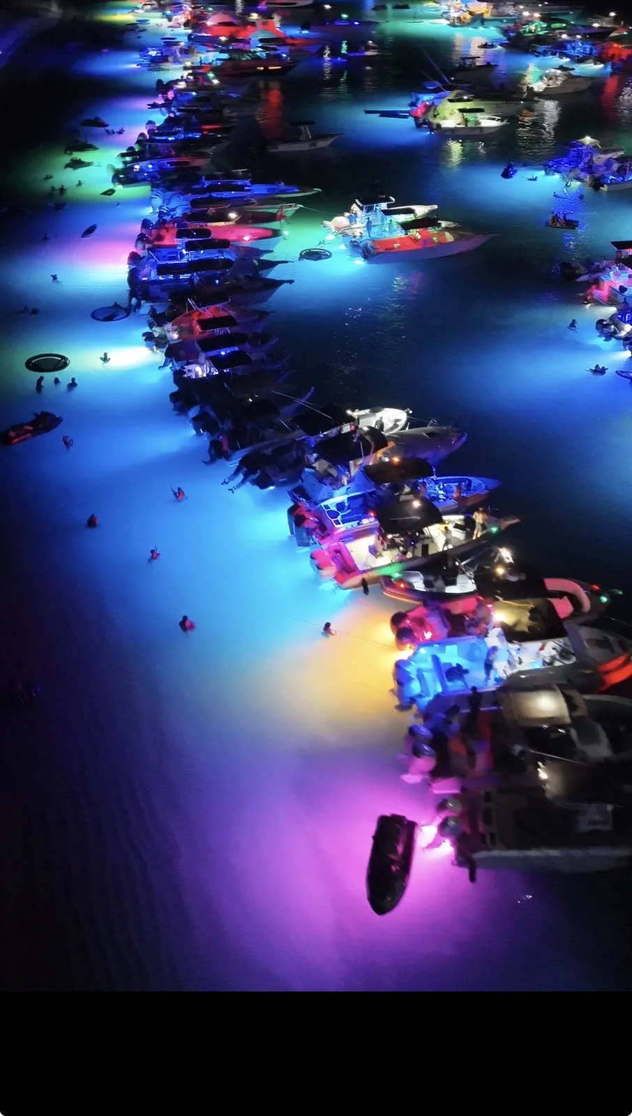 Aerial view of boats rafted up at night off Palomino Island in Puerto Rico with color-changing underwater lights turning the water blue, pink and green