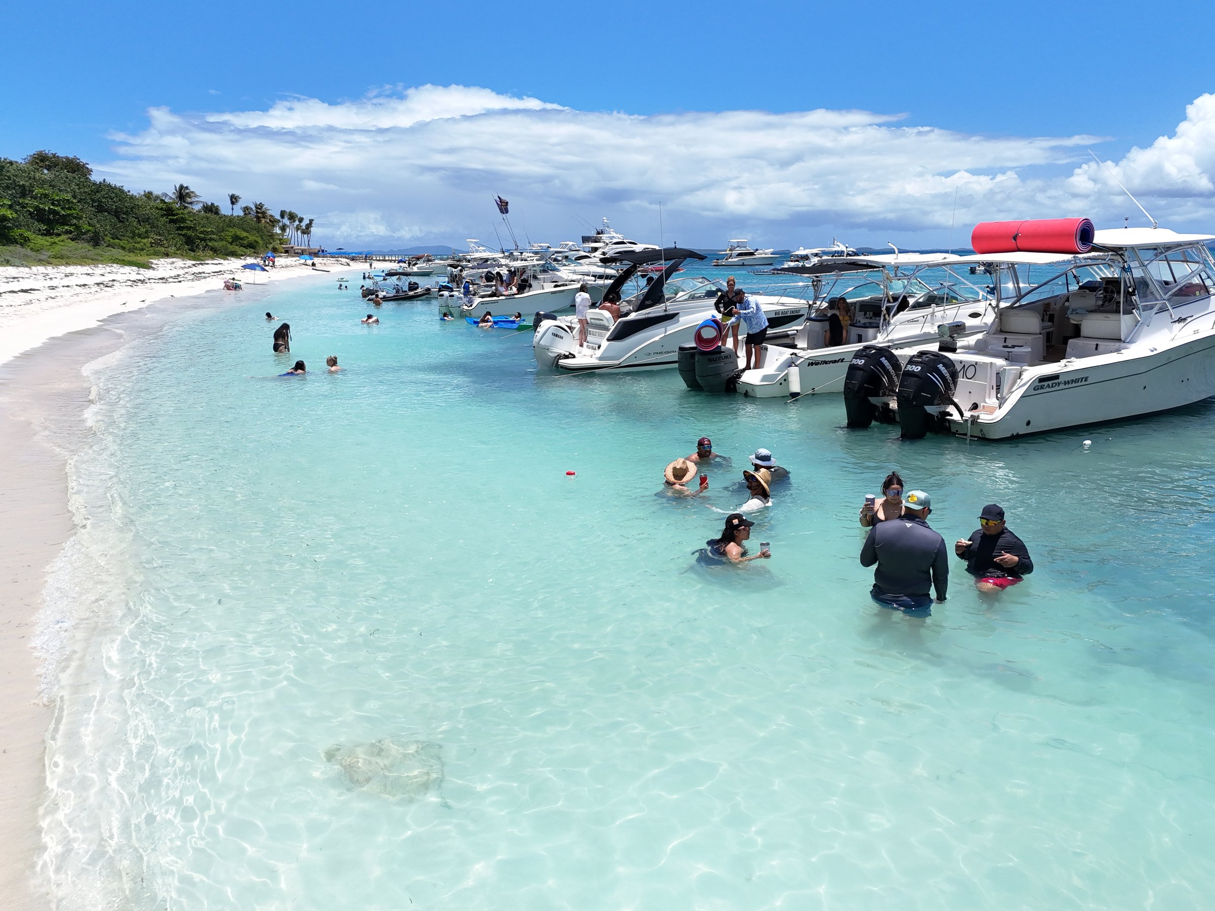 Beach-level view of charter guests in crystal-clear water with multiple yachts anchored at the sandbar